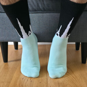 Primer plano de una persona vistiendo calcetines de caña alta bicolor, con fondo negro y siluetas blancas de monumentos del mundo como la Torre Eiffel, y pie en tono verde agua.