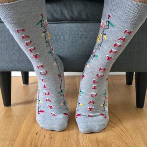 Primer plano de una persona vistiendo calcetines navideños grises con una guirnalda de luces de colores, iconos festivos y el texto vertical "HAPPY HOLIDAYS".