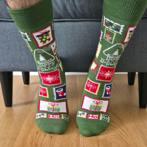 Primer plano de una persona vistiendo calcetines navideños de color verde con estampado de sellos postales festivos, regalos y matasellos "Happy Holidays".